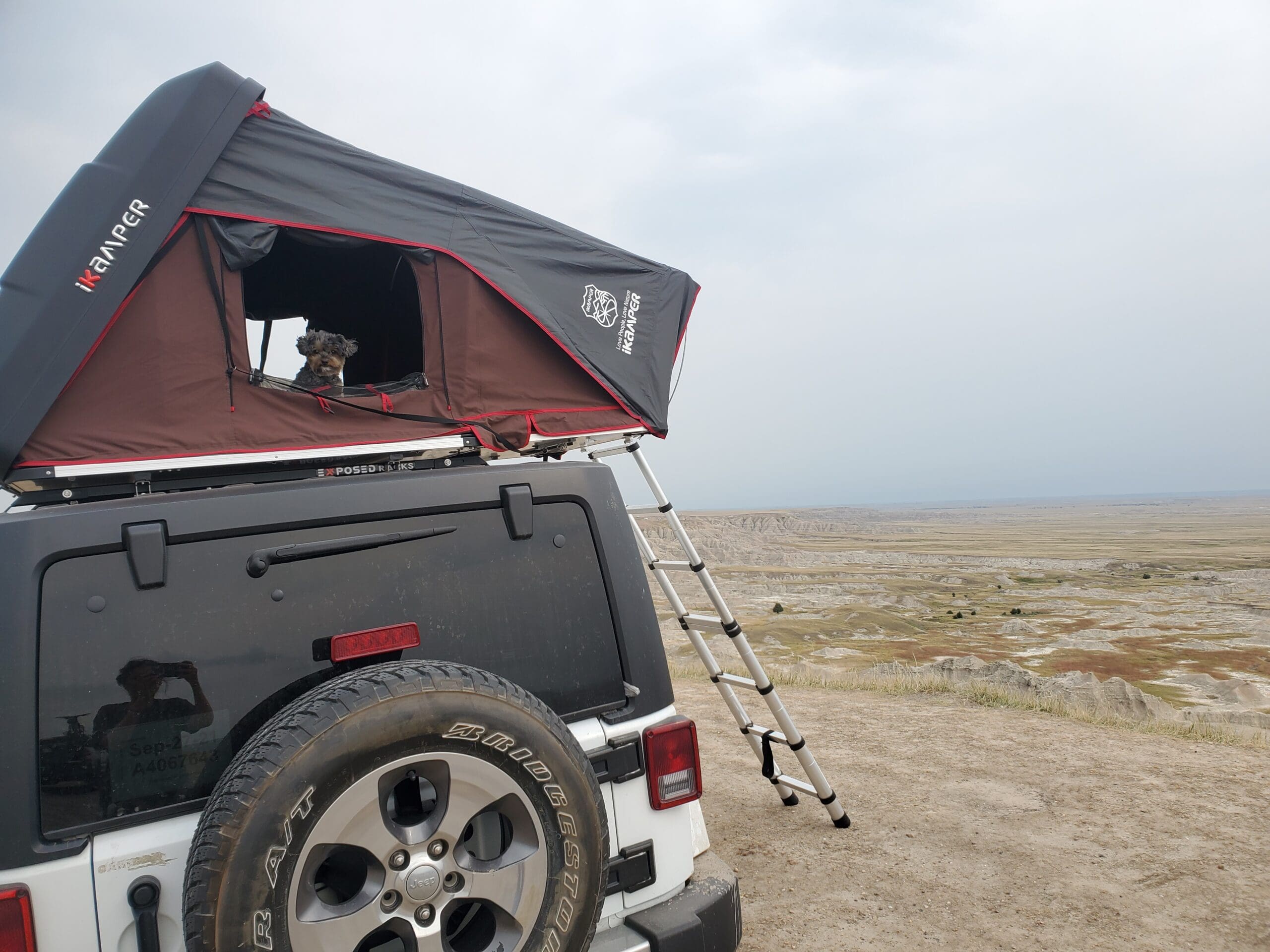 My open Ikamper Mini Rooftop Tent on top of my white Jeep Wrangler on a cliff overlooking the Badlands National Park in South Dakota with my Yorkipoo Dog Loki peering out the open side window at me. His iKamper Review is pretty solid - dogs love iKampers