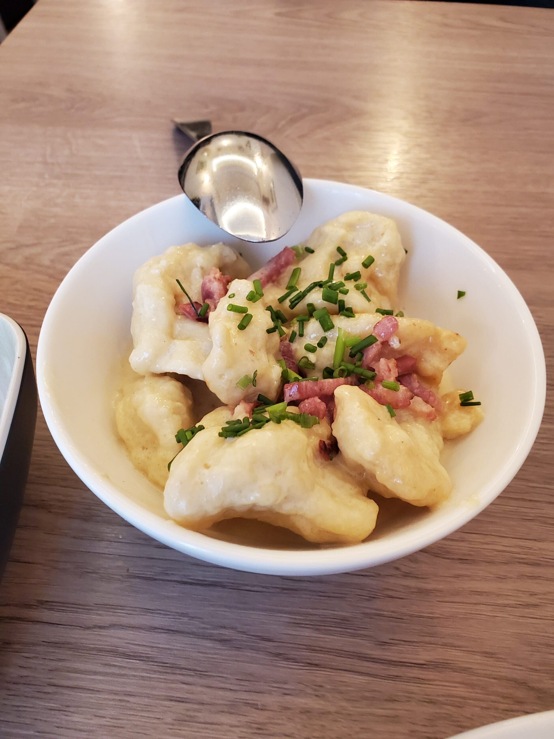 Kniddelen - a white bowl of flour dumplings, sour cream, bacon, and chives with a silver spoon placed on the bowl