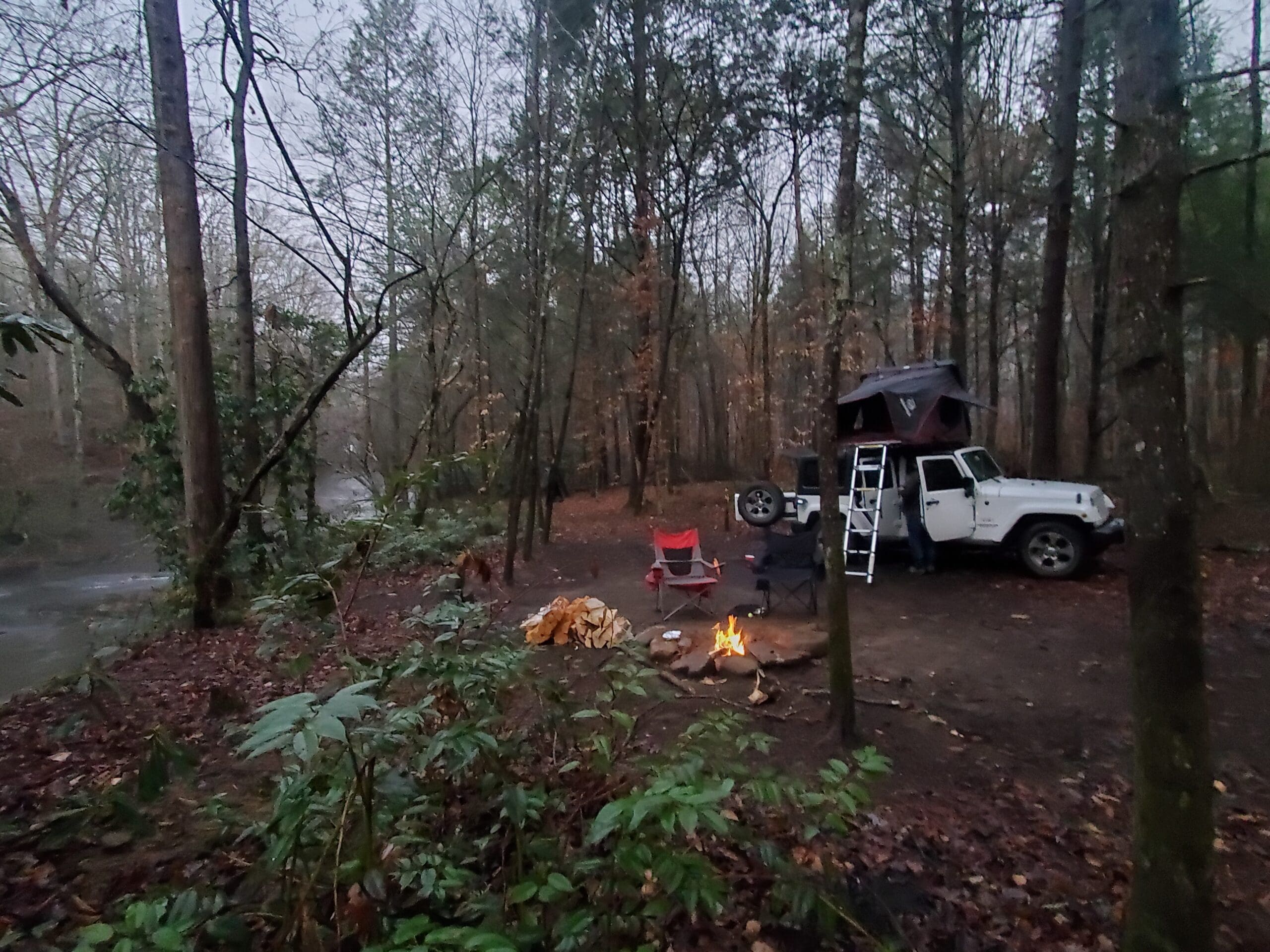 My fully set up iKamper Mini Rooftop Tent at a campsite in the middle of a deciduous forest. Two chairs are in front of a crackling fire, with a full pile of firewood nearby. The Rooftop tent and jeep are behind the chairs overlooking the fire.