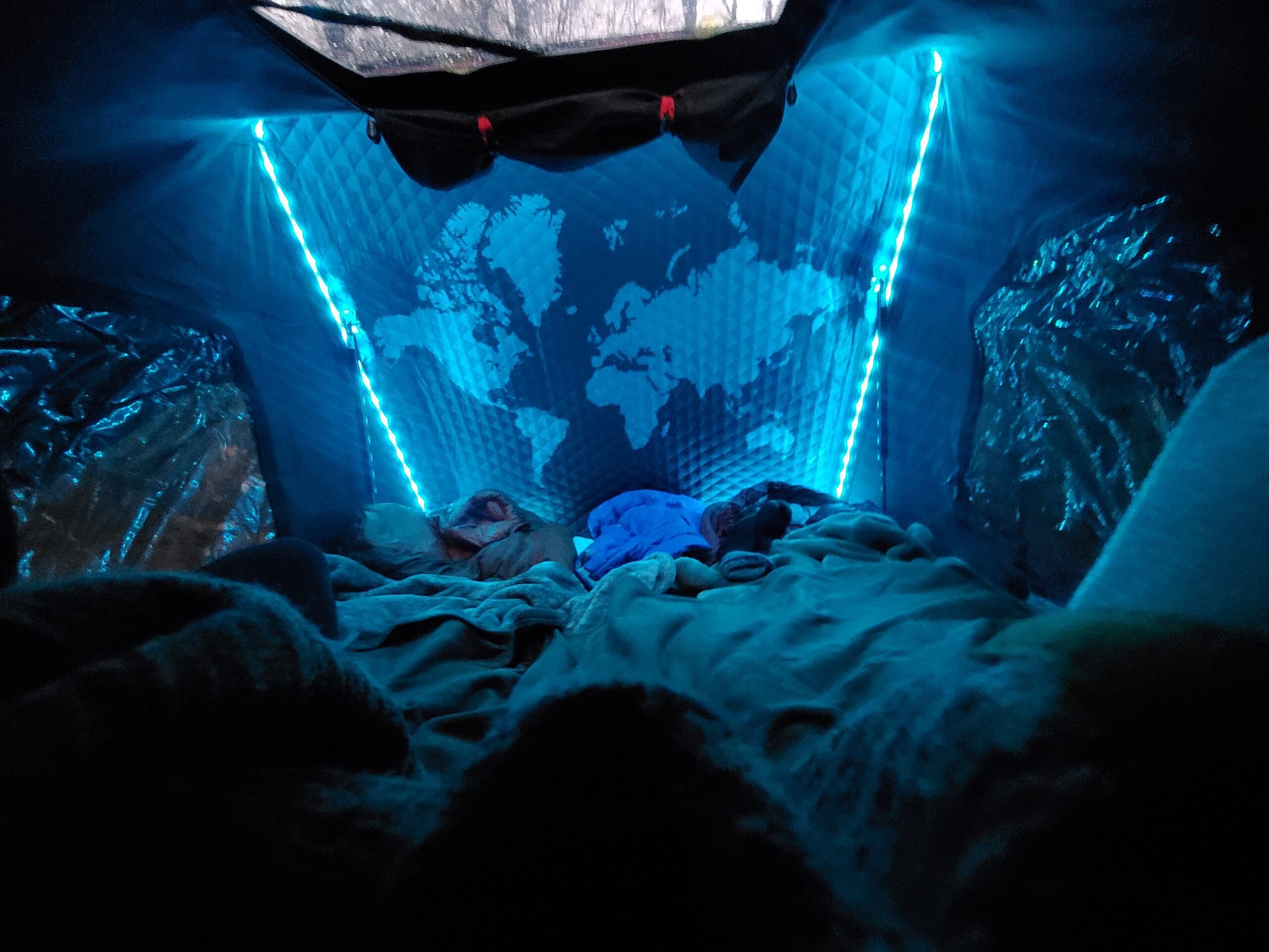 The interior of my iKamper Rooftop Tent Mini. A world map is on the background and the LED Strip Lights are around the perimeter of the map. The lights on and glowing light blue in the background. There are two sleeping bags set up and the skylight at the top of the tent is open. 