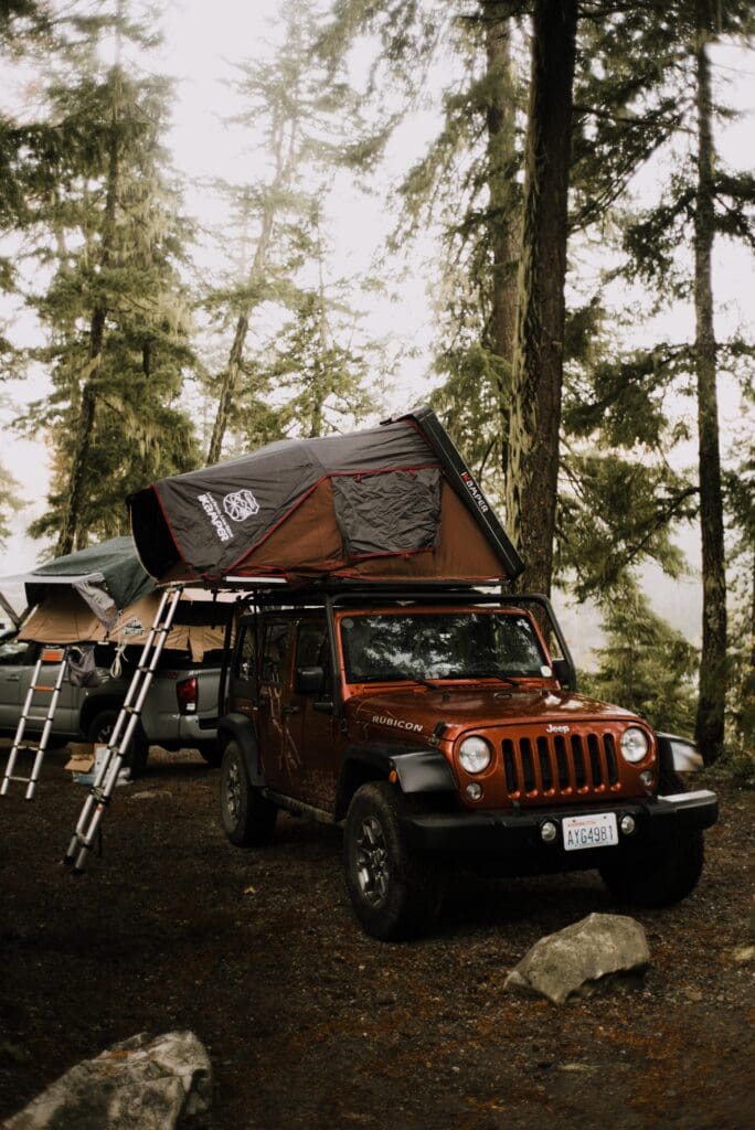 In this iKamper Review, the photo shows an open full size iKamper Rooftoop tent on top of a burnt orange jeep in a fir tree forest at a campsite. 