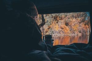 A man drinking coffee in the morning looking out of a rooftop tent over a calm lake during autumn.