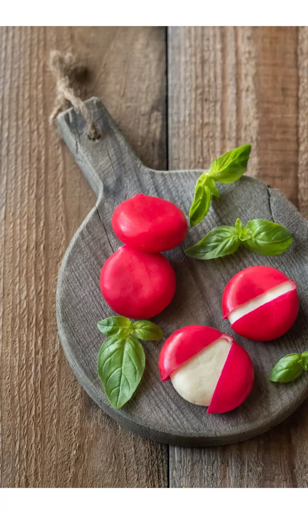 Babybel cheeses wrapped in red wax on a wooden board with basil leaves sprinkled around