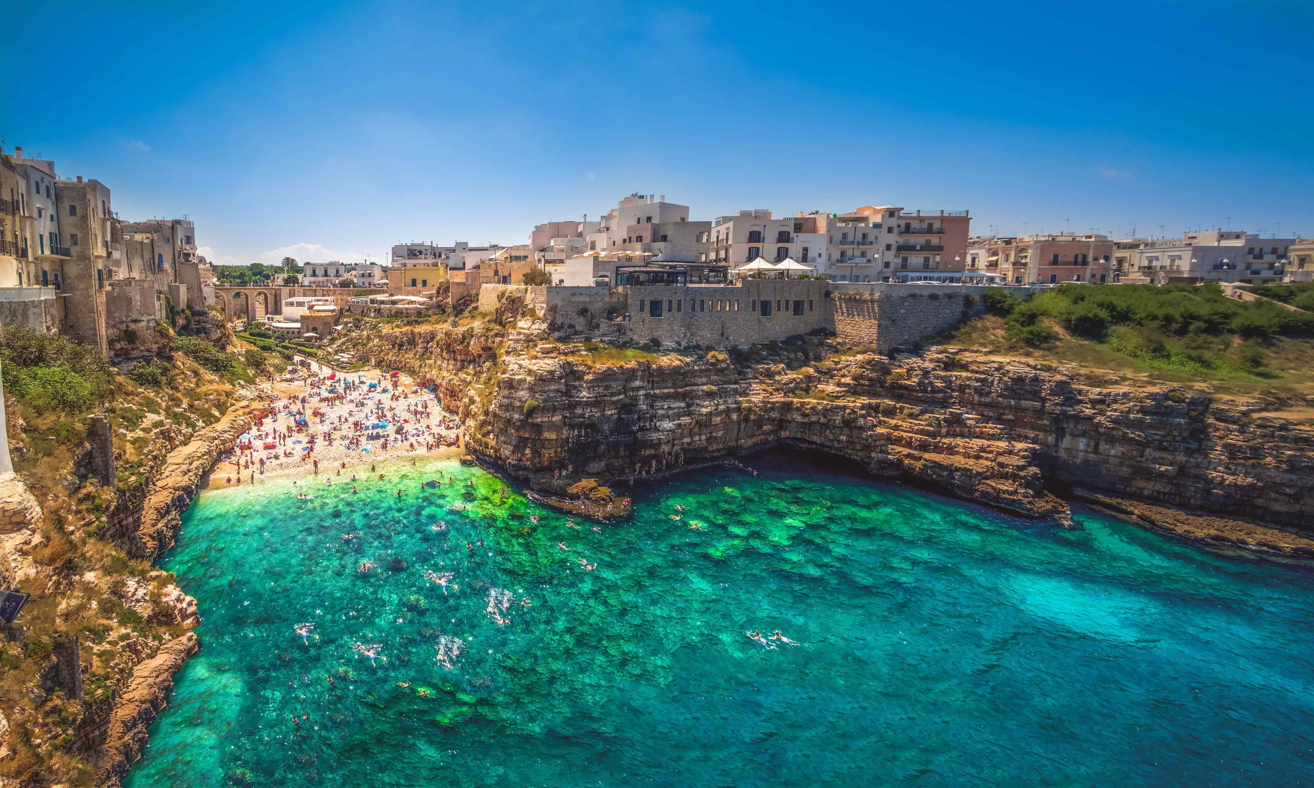 Lama Monachile is one of the best beaches in Puglia, Italy. This photo is of a thin, golden sand beach nestled amongst tall fliffs and old school Italian apartments. The water is very blue and rocks are visible at the bottom.