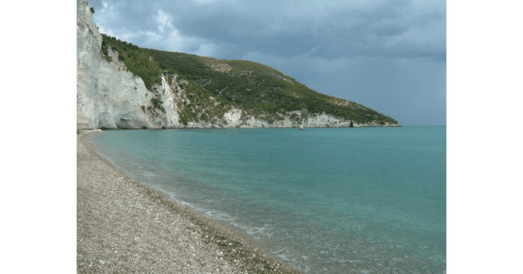 This is Spiaggia di Vignanotica Beach. A pebble beach with white cliffs topped by green trees. The sky is stormy.