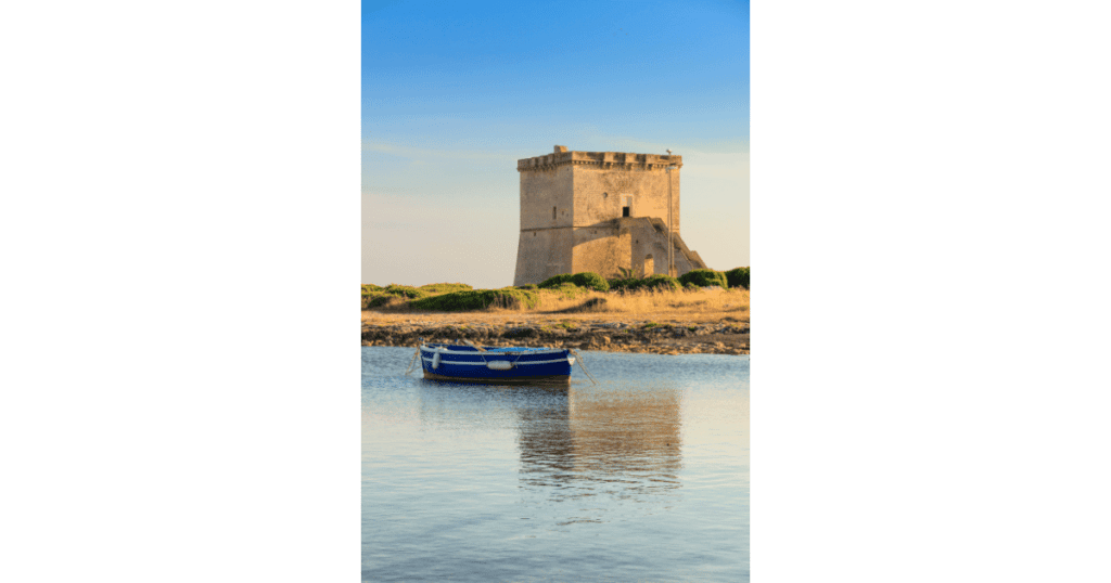 A historic fort tower against a blue sky and in the foreground, tranquil waters and a small blue fishing boat floating serenely.